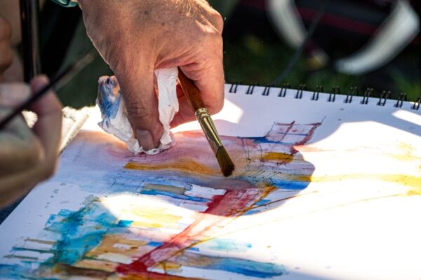Close-up of a mature artist's hands actively painting a watercolor sketch with a brush, working on a spiral-bound sketchbook in bright natural daylight. Vibrant blues, yellows, and reds are visible on the white paper, with the artist's hand showing age spots and wearing a simple ring. The outdoor setting is blurred in the background, suggesting a plein air sketching session, with warm golden sunlight illuminating the work surface.